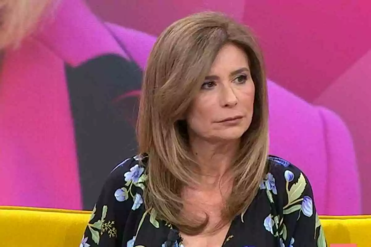 Gema López in a floral print blouse sitting on a yellow sofa with a pink background.