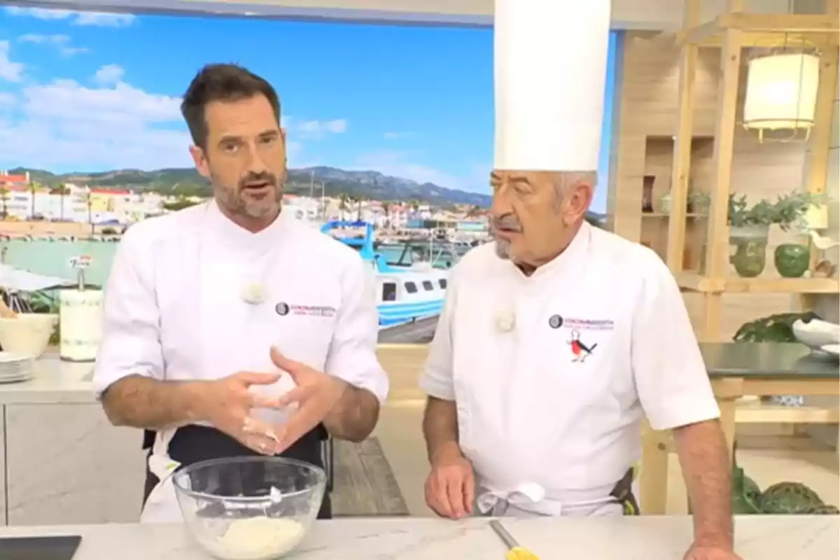 Karlos y Joseba Arguiñano con uniforme blanco conversan en una cocina moderna mientras Joseba mezcla ingredientes en un bol de cristal y Karlos lleva un gorro alto de chef Karlos y Joseba Arguiñano con uniforme blanco conversan en una cocina moderna mientras Joseba mezcla ingredientes en un bol de cristal y Karlos lleva un gorro alto de chef
