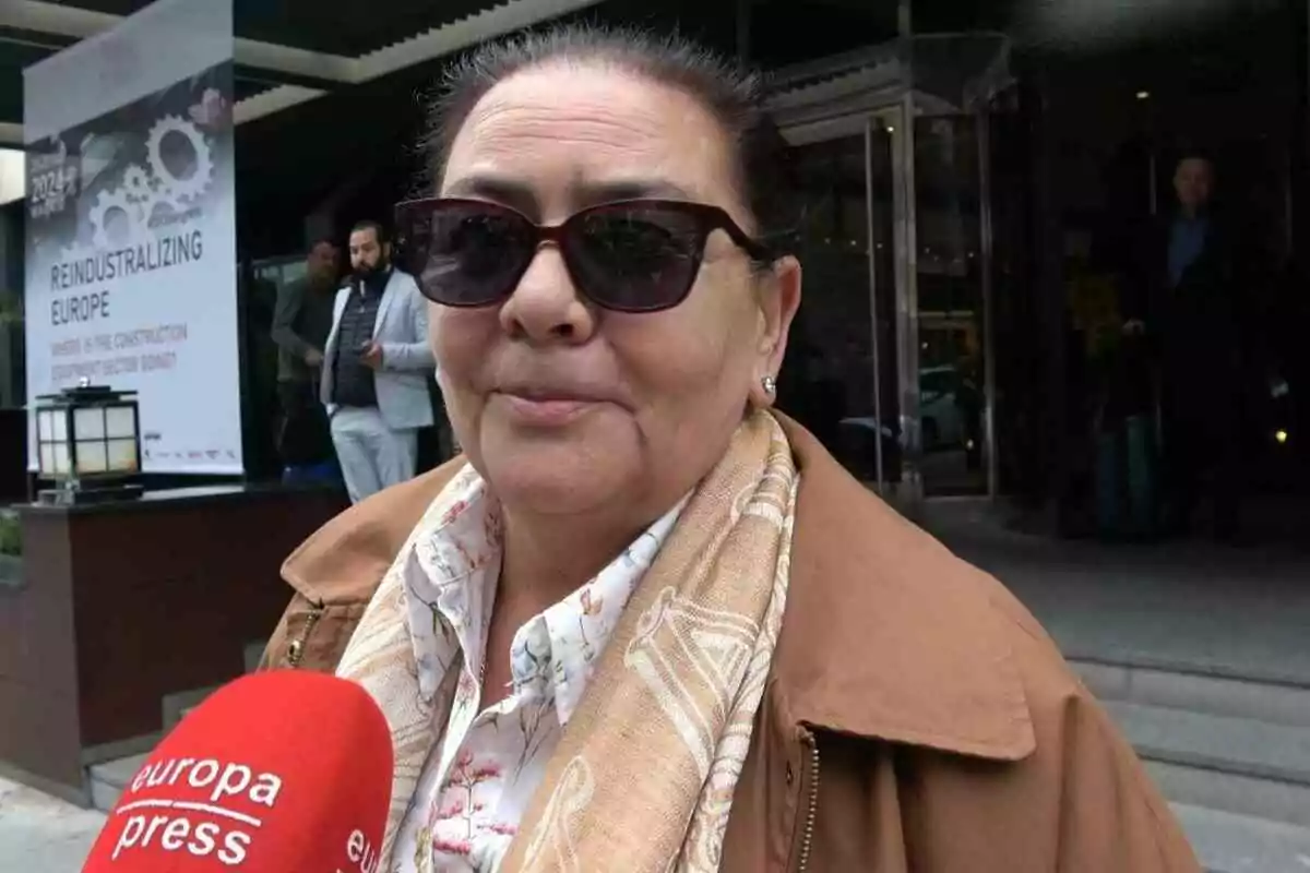 María del Monte with sunglasses and a scarf is being interviewed by a Europa Press microphone in front of a building with a sign that says 