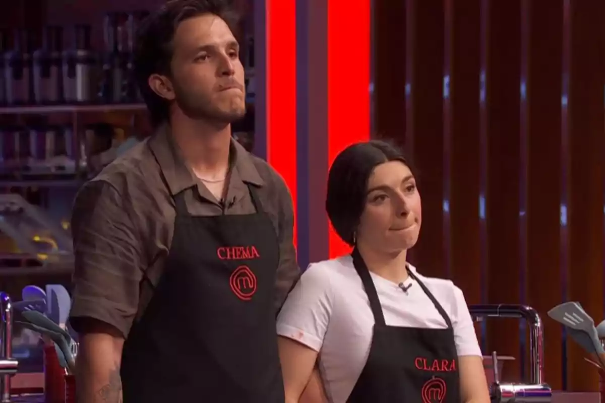 Clara y Chema, concursantes de 'Masterchef', con delantales negros y expresión seria en una cocina iluminada con luces rojas. Clara y Chema, concursantes de 'Masterchef', con delantales negros y expresión seria en una cocina iluminada con luces rojas.