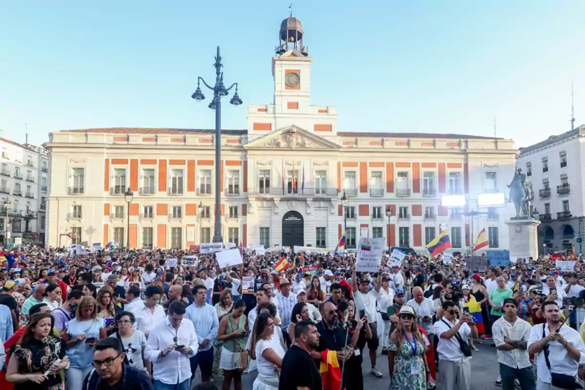 Una multitud de personas se reúne frente a un edificio histórico en una plaza, algunas sosteniendo pancartas y banderas.