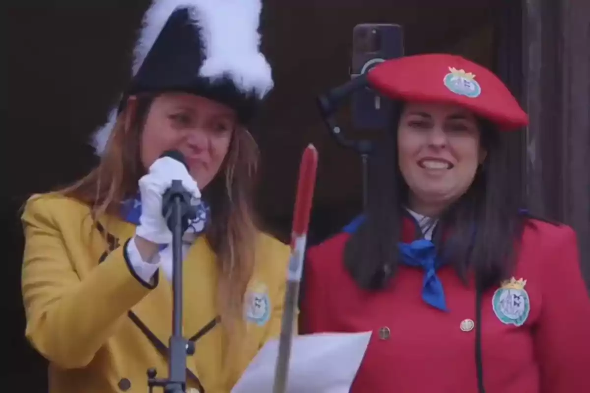 Dos mujeres vestidas con uniformes coloridos, una de ellas con chaqueta amarilla y sombrero negro con plumas blancas, hablando en un micrófono, mientras la otra, con chaqueta roja y boina roja, sonríe.