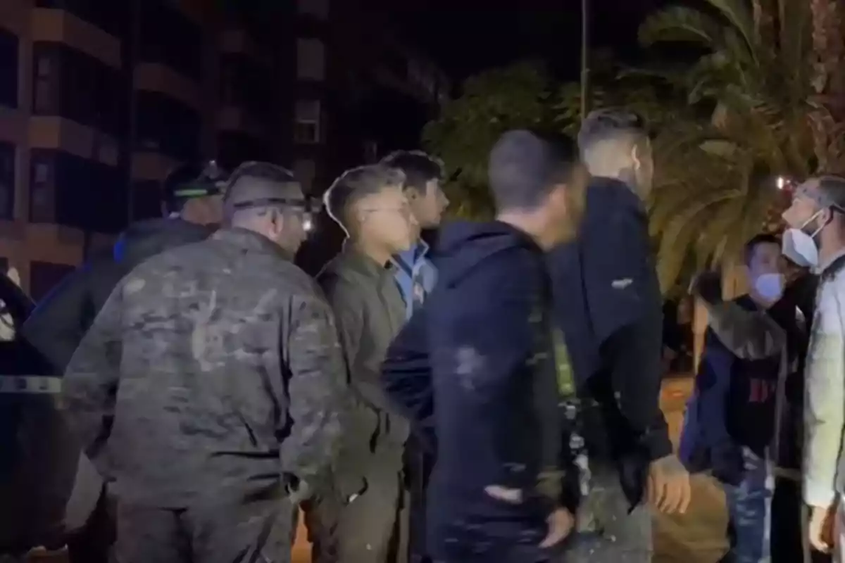 Un grupo de personas, algunas con uniforme militar, se encuentran reunidas en una calle por la noche. Un grupo de personas, algunas con uniforme militar, se encuentran reunidas en una calle por la noche.