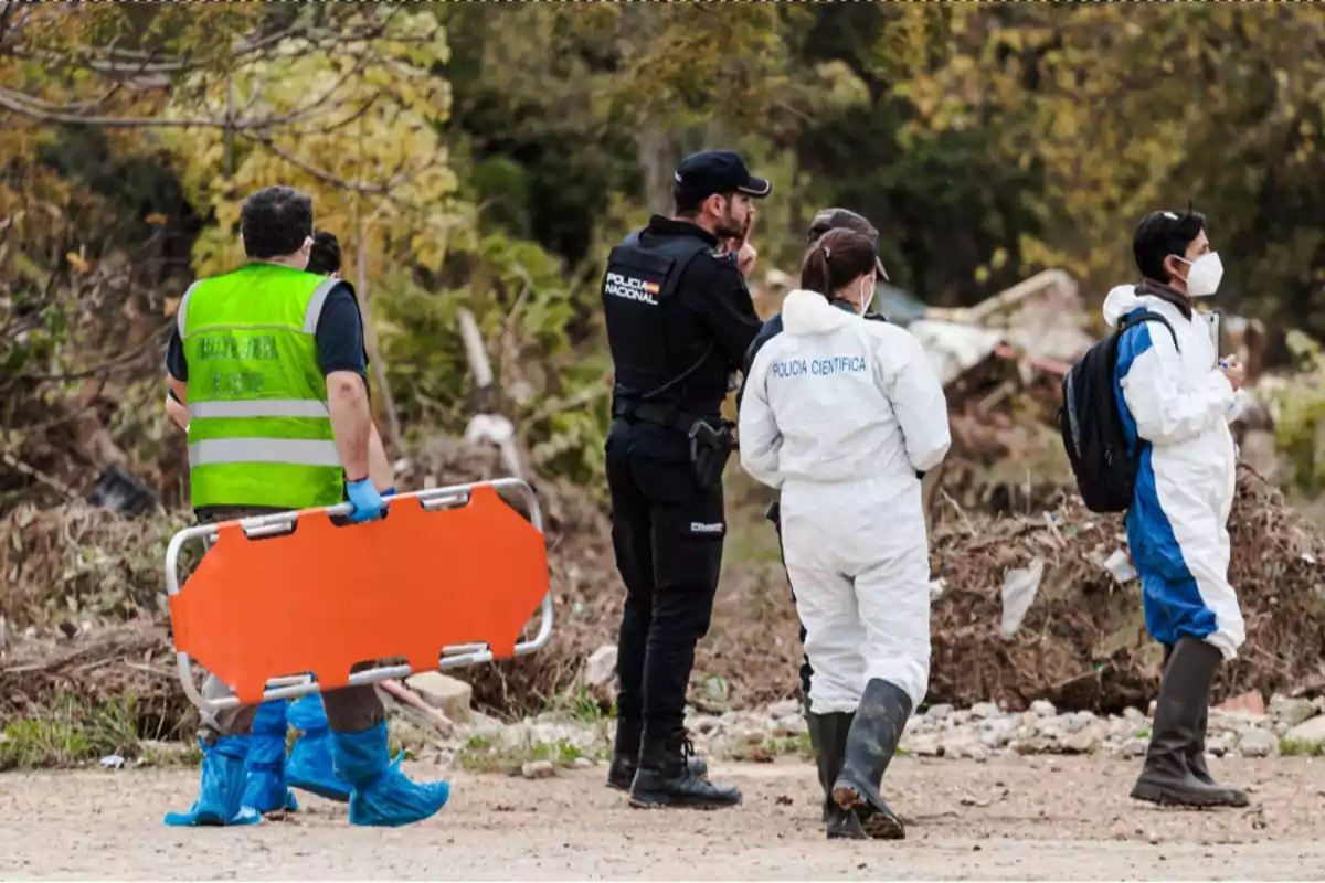 Un grupo de personas con equipo de protección y uniformes de policía científica se encuentra en un área al aire libre, una de ellas lleva una camilla naranja.