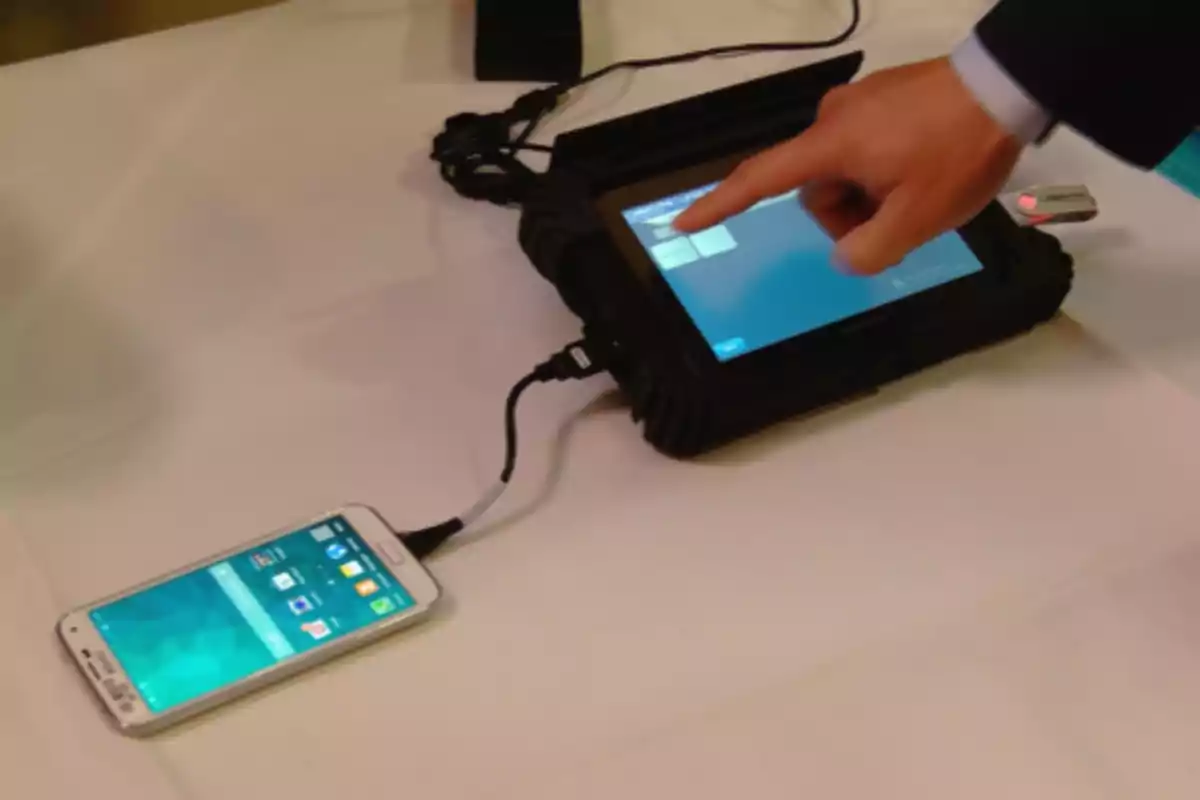 A smartphone connected to an electronic device via a USB cable while a person interacts with the device's touchscreen.