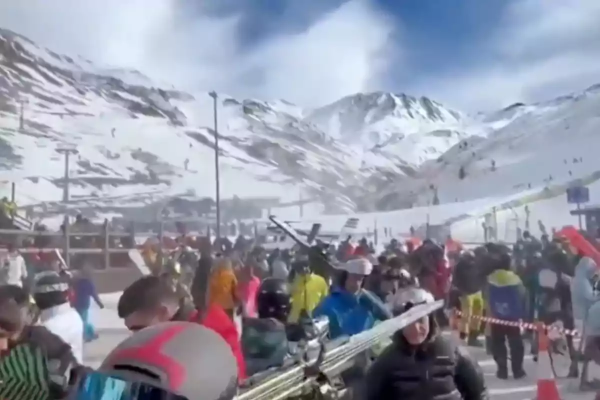 Personas con equipo de esquí en una estación de esquí concurrida con montañas nevadas al fondo.