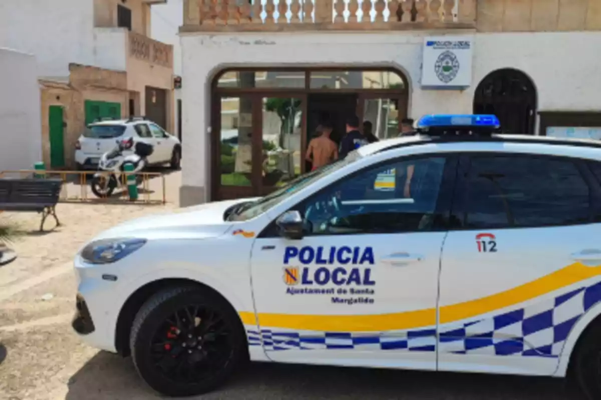 Un coche de la policía local estacionado frente a un edificio con personas en la entrada.