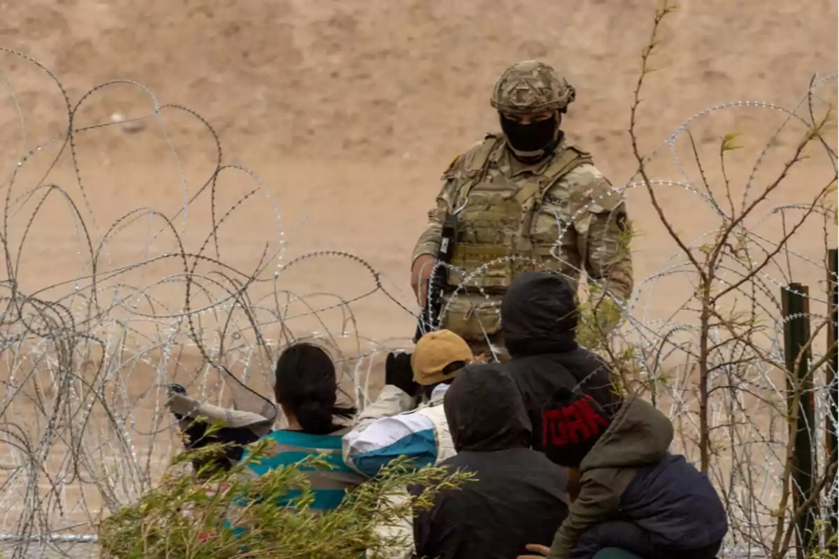 Un soldado en uniforme militar observa a un grupo de personas detrás de una cerca de alambre de púas en un entorno desértico.