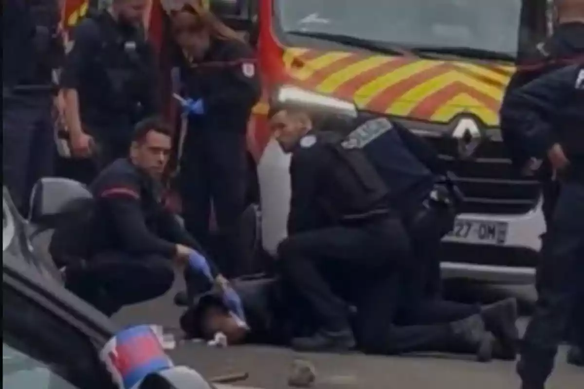 Un grupo de policías atiende a una persona en el suelo frente a un vehículo de emergencia con franjas amarillas y rojas.