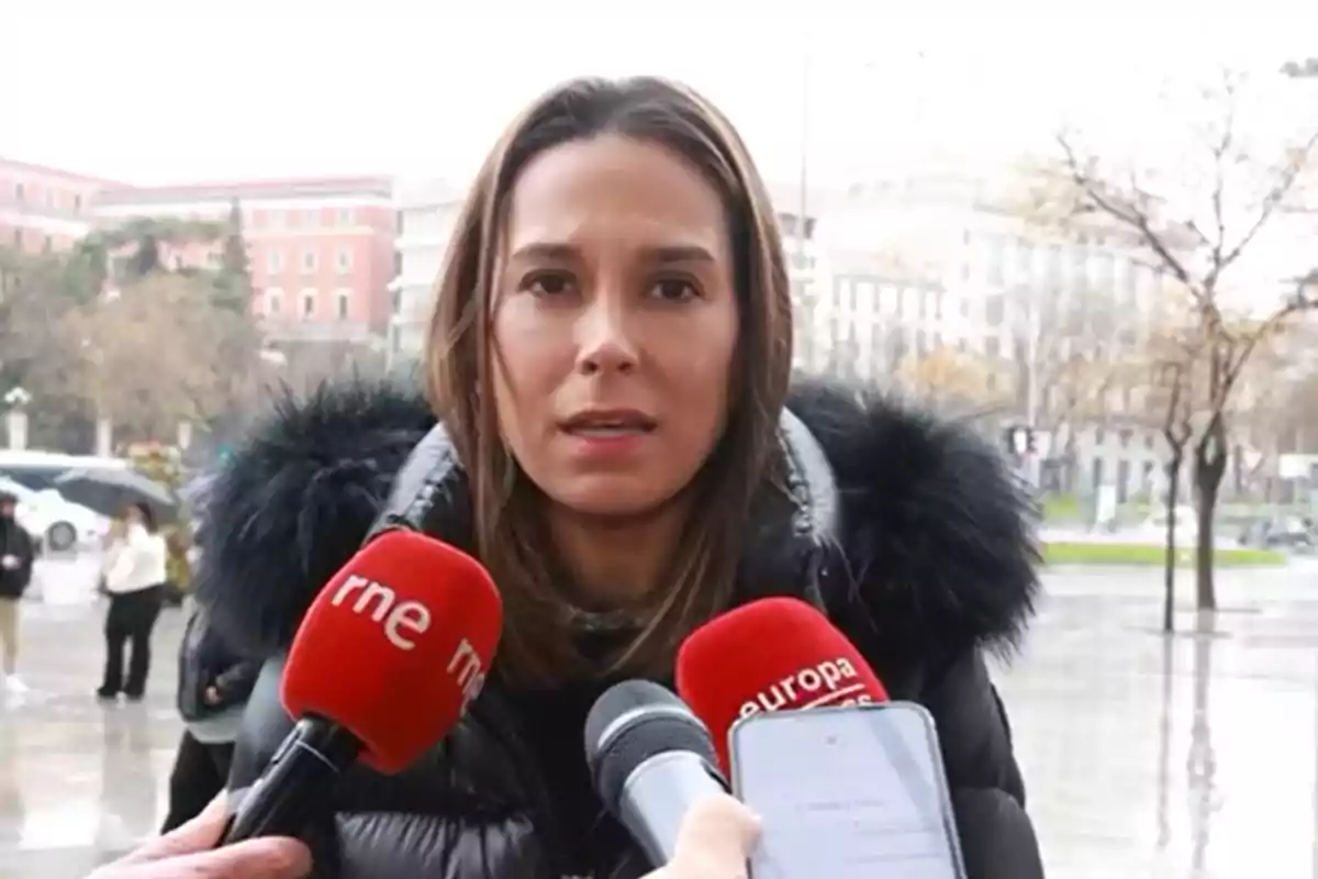 A woman in a winter jacket is being interviewed by various media outlets in an urban setting.