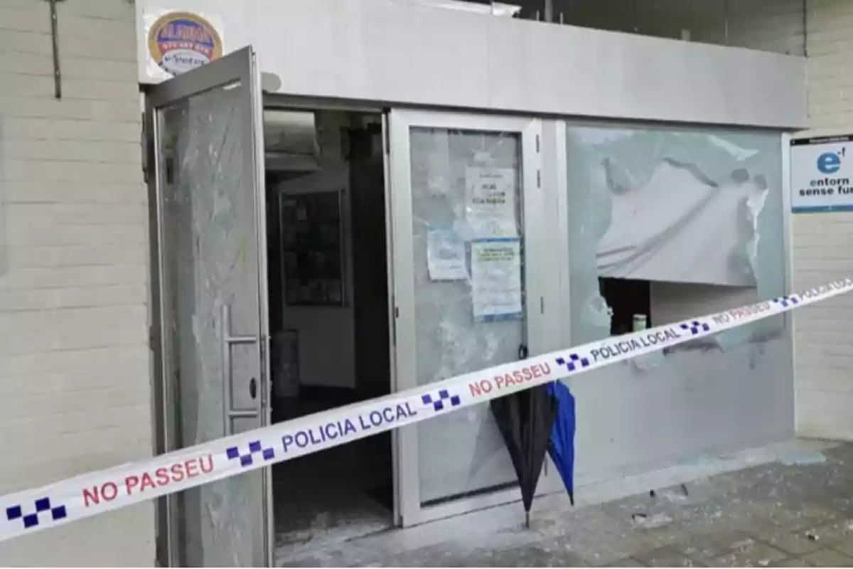 Una puerta de vidrio rota con cinta de la policía local que dice "NO PASSEU" bloqueando el acceso.