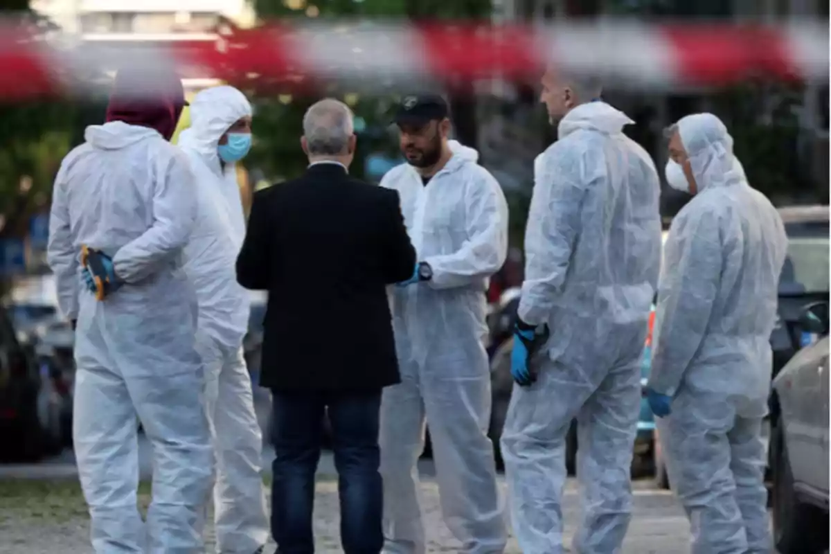 Un grupo de personas con trajes de protección blanca y mascarillas conversa en una calle, mientras una cinta de seguridad roja y blanca se ve desenfocada en primer plano. Un grupo de personas con trajes de protección blanca y mascarillas conversa en una calle, mientras una cinta de seguridad roja y blanca se ve desenfocada en primer plano.