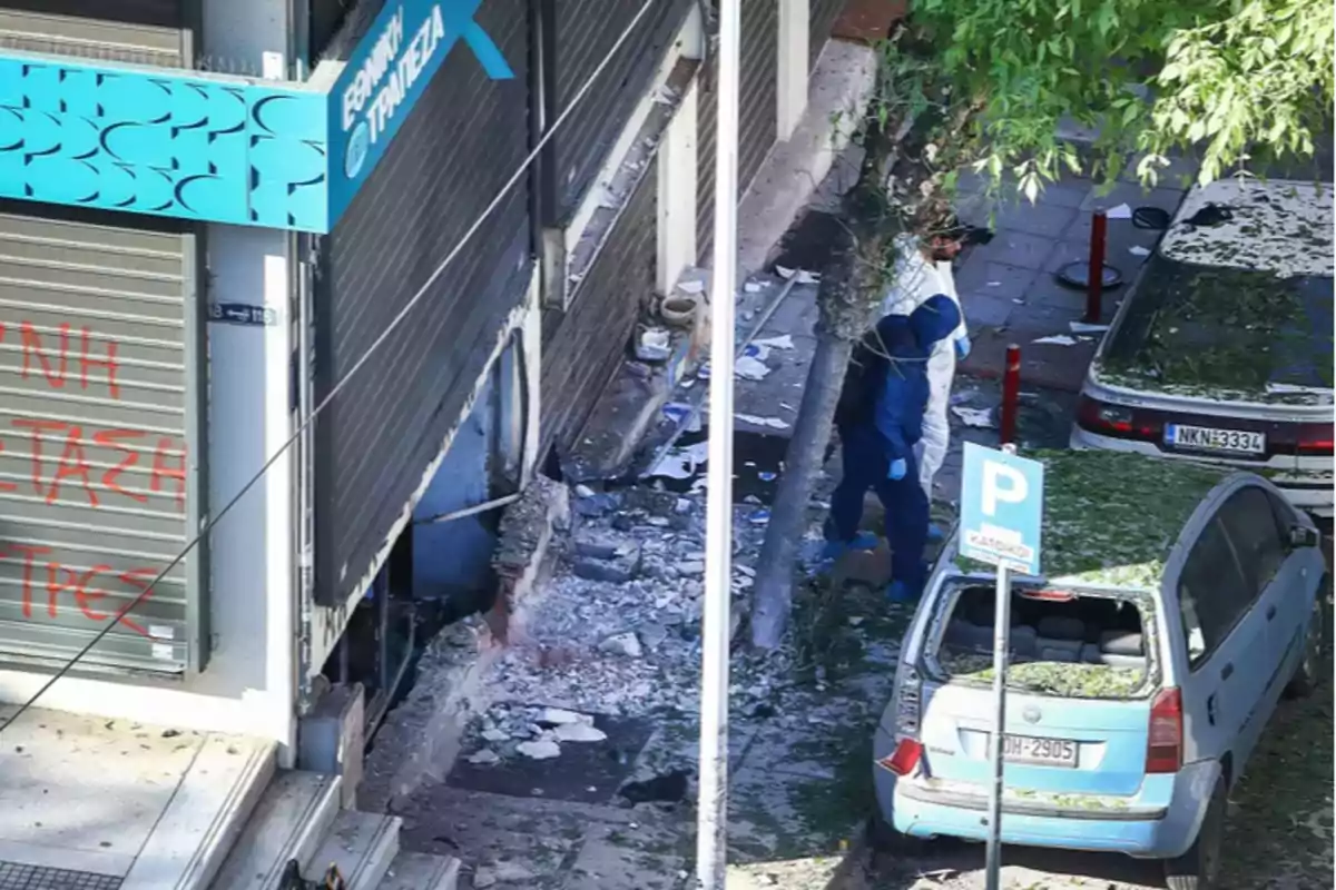 Un edificio con daños visibles en la fachada y escombros en el suelo, con dos personas vestidas con trajes de protección caminando cerca de un coche estacionado.