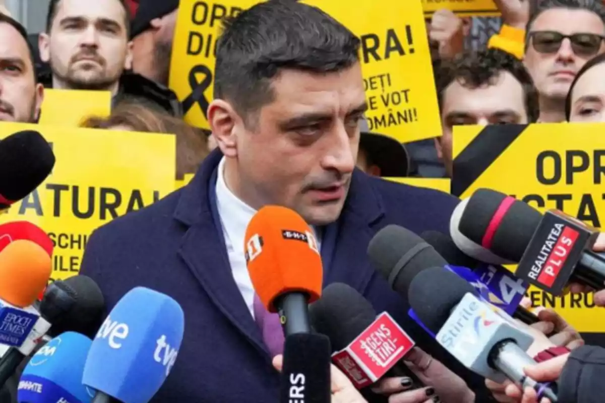 Un hombre rodeado de micrófonos de prensa mientras personas sostienen carteles amarillos de protesta en el fondo. Un hombre rodeado de micrófonos de prensa mientras personas sostienen carteles amarillos de protesta en el fondo.