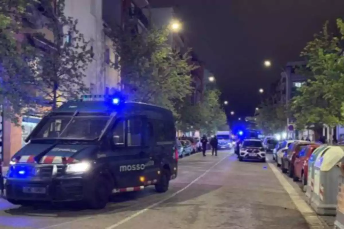 Una calle de noche con vehículos policiales y luces azules encendidas, rodeada de árboles y edificios.