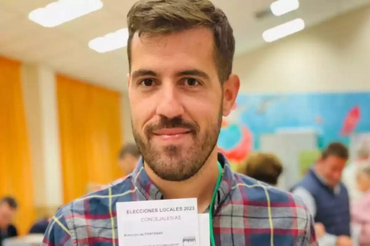 Un hombre con camisa de cuadros sostiene un documento en un lugar con varias personas al fondo. Un hombre con camisa de cuadros sostiene un documento en un lugar con varias personas al fondo.