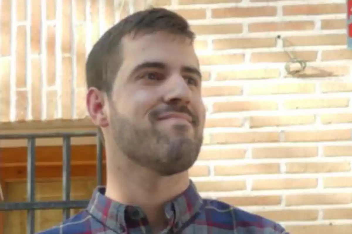 Un hombre con barba y camisa a cuadros sonríe ligeramente mientras está de pie frente a una pared de ladrillos.
