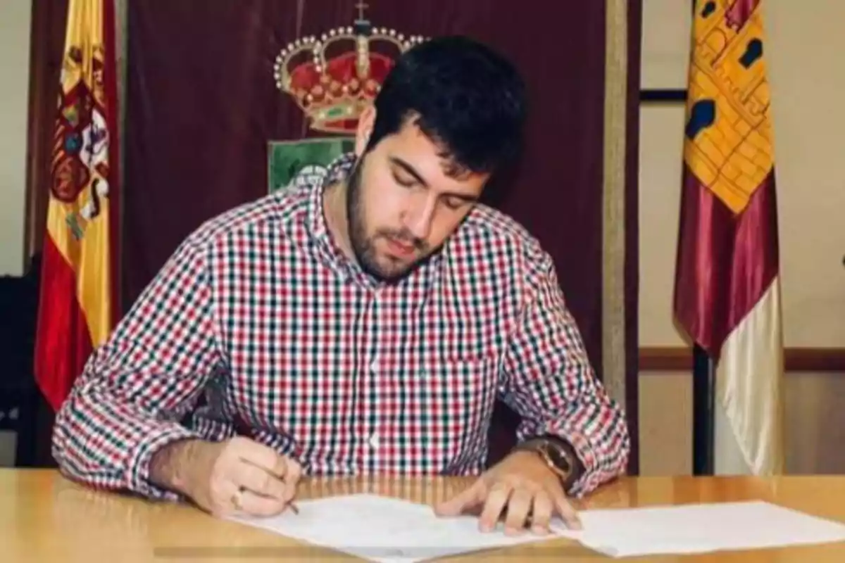 Un hombre con camisa de cuadros está sentado en una mesa firmando documentos, con banderas y un escudo en el fondo. Un hombre con camisa de cuadros está sentado en una mesa firmando documentos, con banderas y un escudo en el fondo.