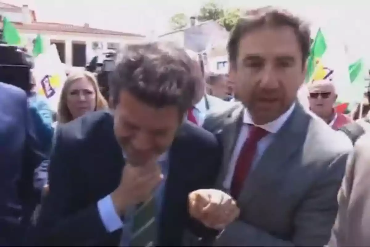 Un grupo de personas en un evento al aire libre, donde dos hombres en el centro parecen interactuar de manera amistosa, rodeados de otras personas y banderas en el fondo. Un grupo de personas en un evento al aire libre, donde dos hombres en el centro parecen interactuar de manera amistosa, rodeados de otras personas y banderas en el fondo.