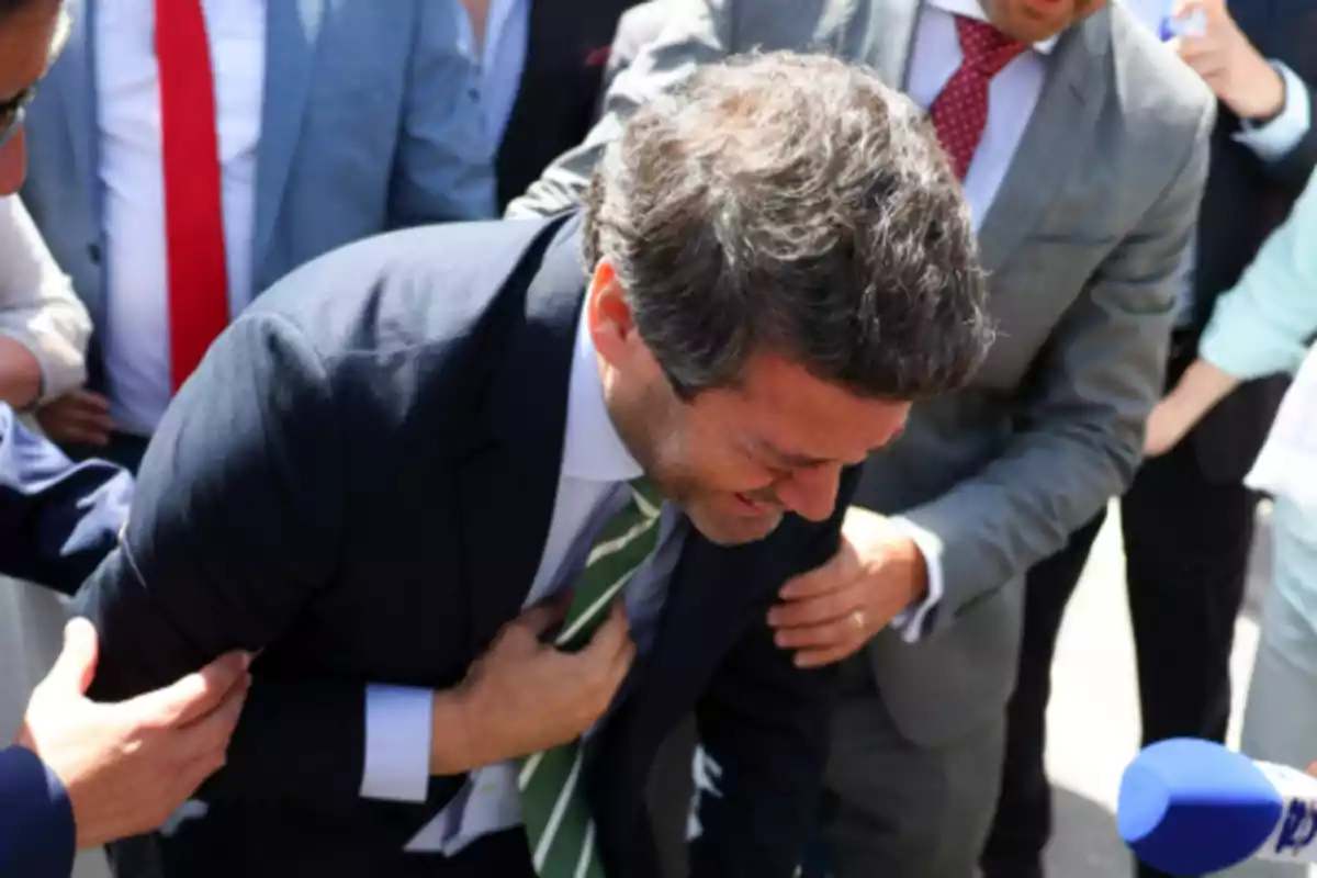 Un hombre con traje oscuro y corbata verde se inclina hacia adelante mientras es asistido por otras personas a su alrededor.