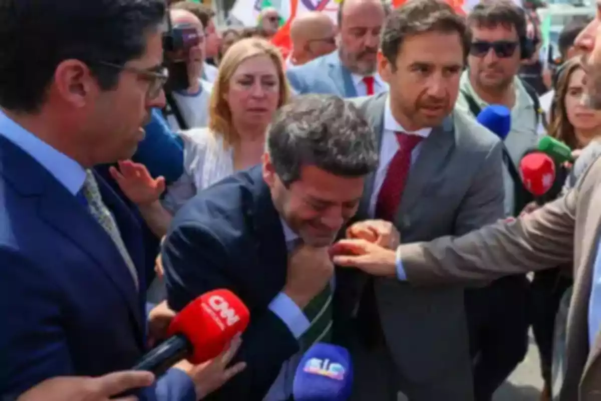 Un grupo de personas rodea a un hombre que parece estar en una situación tensa mientras varios micrófonos de medios de comunicación están presentes. Un grupo de personas rodea a un hombre que parece estar en una situación tensa mientras varios micrófonos de medios de comunicación están presentes.