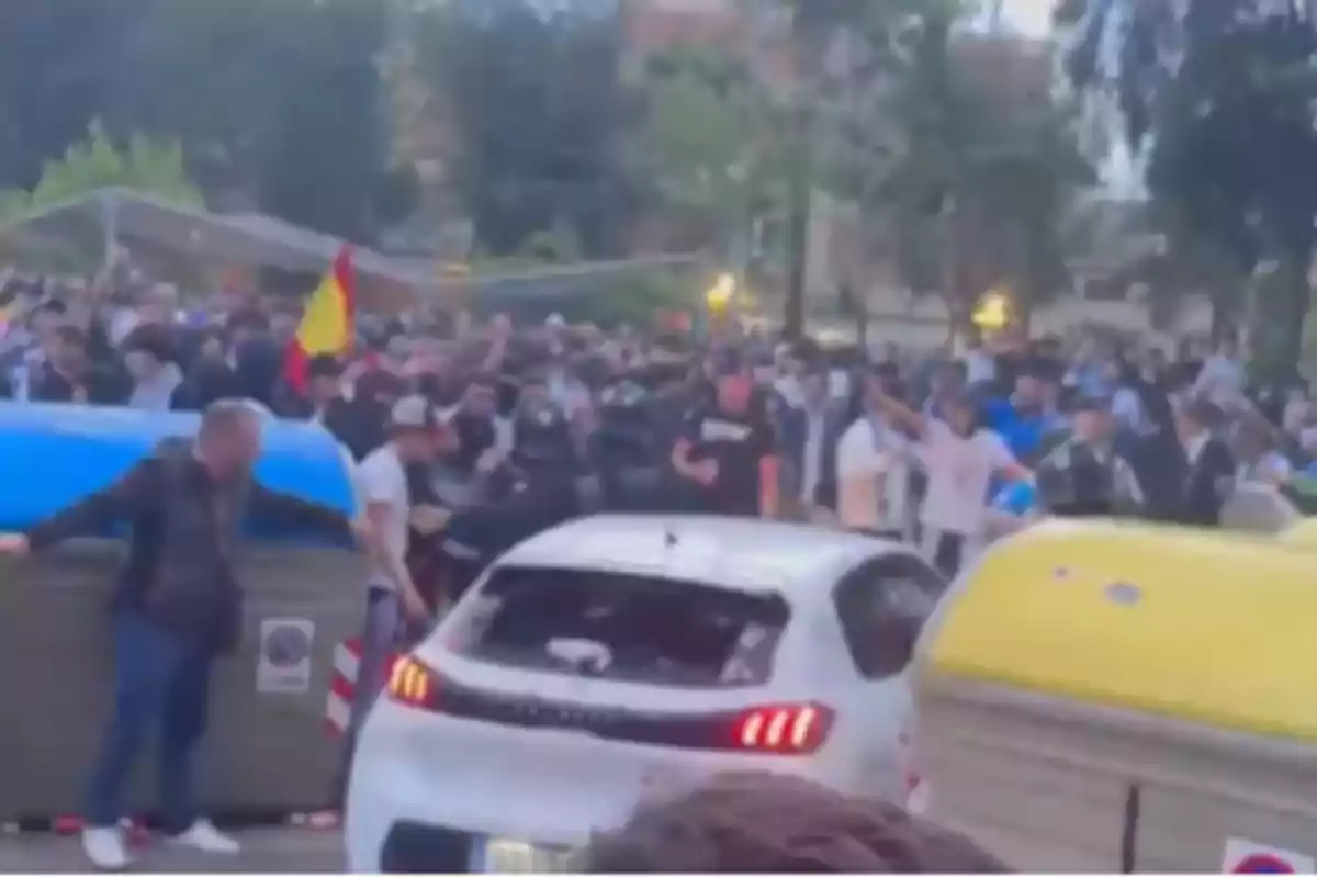 Una multitud se reúne alrededor de un coche blanco en una calle, con contenedores de basura a los lados y una bandera visible entre la gente. Una multitud se reúne alrededor de un coche blanco en una calle, con contenedores de basura a los lados y una bandera visible entre la gente.