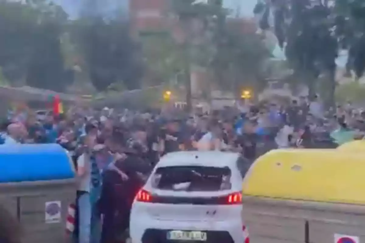 Una multitud rodea un coche blanco en una calle con contenedores de basura en primer plano.
