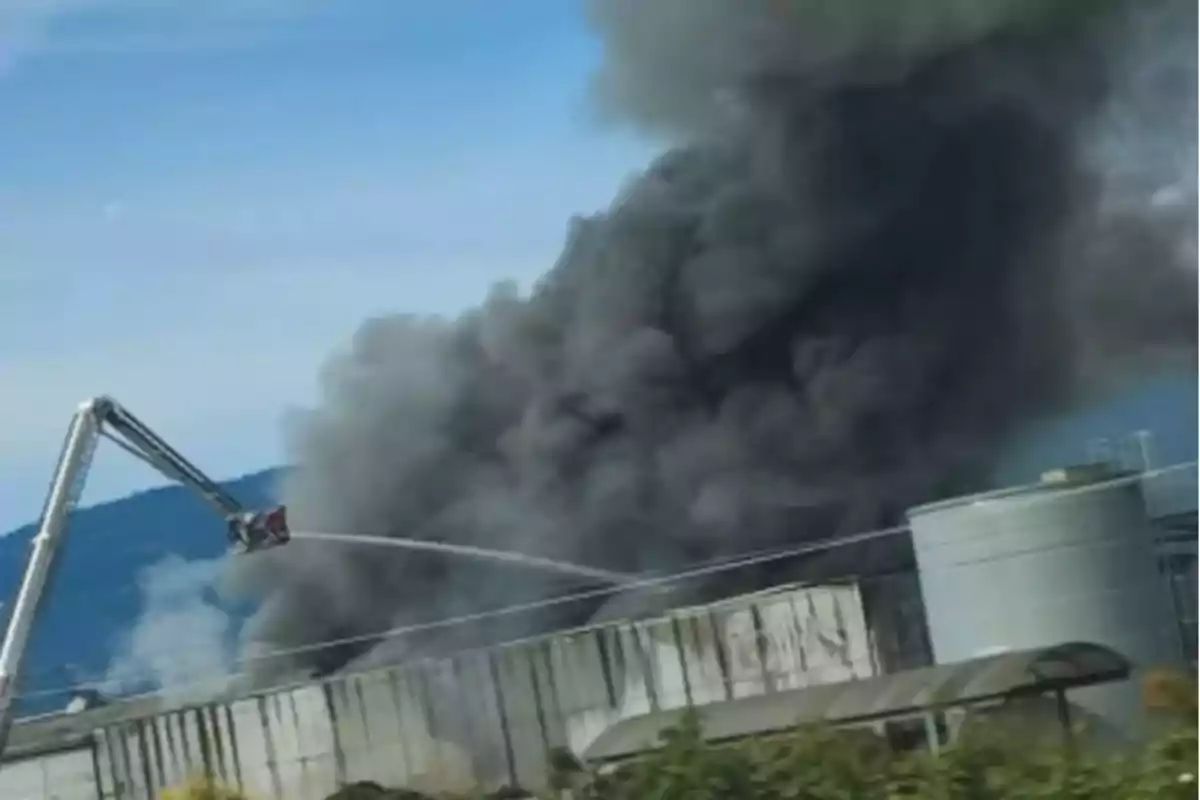 Una columna de humo negro se eleva desde un edificio industrial mientras un camión de bomberos con una escalera extiende una manguera para combatir el incendio. Una columna de humo negro se eleva desde un edificio industrial mientras un camión de bomberos con una escalera extiende una manguera para combatir el incendio.
