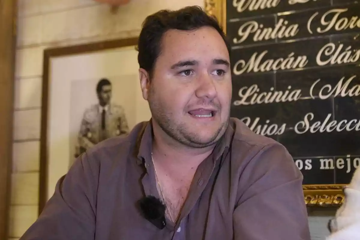 Un hombre con camisa oscura habla frente a un fondo con una pizarra escrita y una fotografía enmarcada. Un hombre con camisa oscura habla frente a un fondo con una pizarra escrita y una fotografía enmarcada.