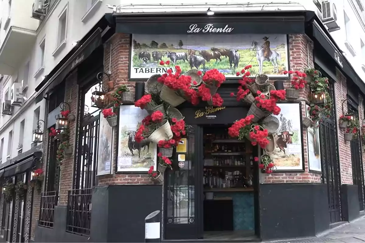 Fachada de una taberna decorada con flores rojas y cestas, con murales de toros y un jinete en las paredes de ladrillo. Fachada de una taberna decorada con flores rojas y cestas, con murales de toros y un jinete en las paredes de ladrillo.