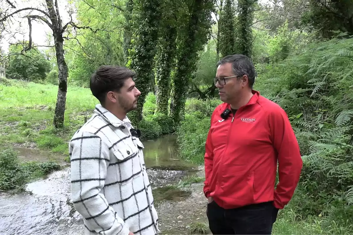 Dos personas conversan junto a un arroyo en un entorno natural rodeado de árboles y vegetación. Dos personas conversan junto a un arroyo en un entorno natural rodeado de árboles y vegetación.