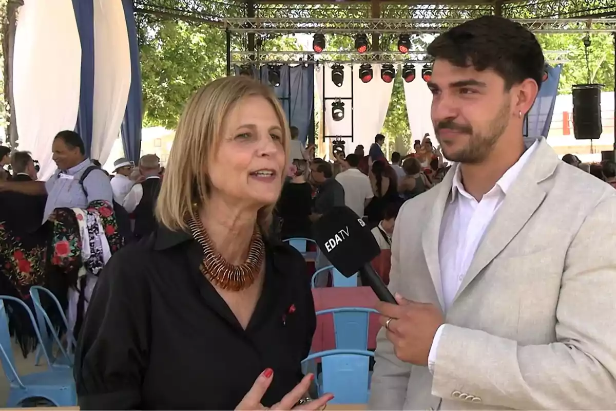 Dos personas están siendo entrevistadas en un evento al aire libre con un micrófono de EDA TV, rodeadas de un grupo de personas y decoraciones festivas. Dos personas están siendo entrevistadas en un evento al aire libre con un micrófono de EDA TV, rodeadas de un grupo de personas y decoraciones festivas.
