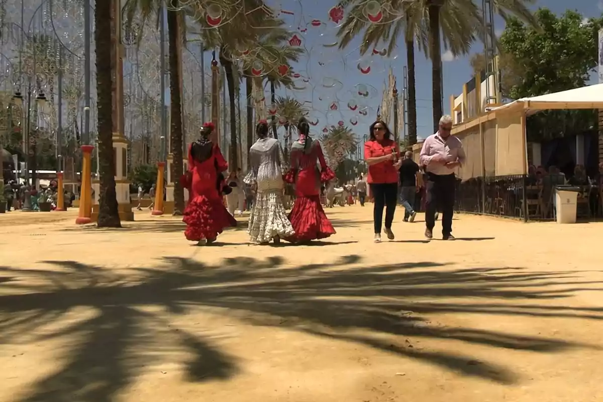 Personas caminando por un paseo adornado con luces y palmeras durante una feria, algunas vestidas con trajes tradicionales. Personas caminando por un paseo adornado con luces y palmeras durante una feria, algunas vestidas con trajes tradicionales.