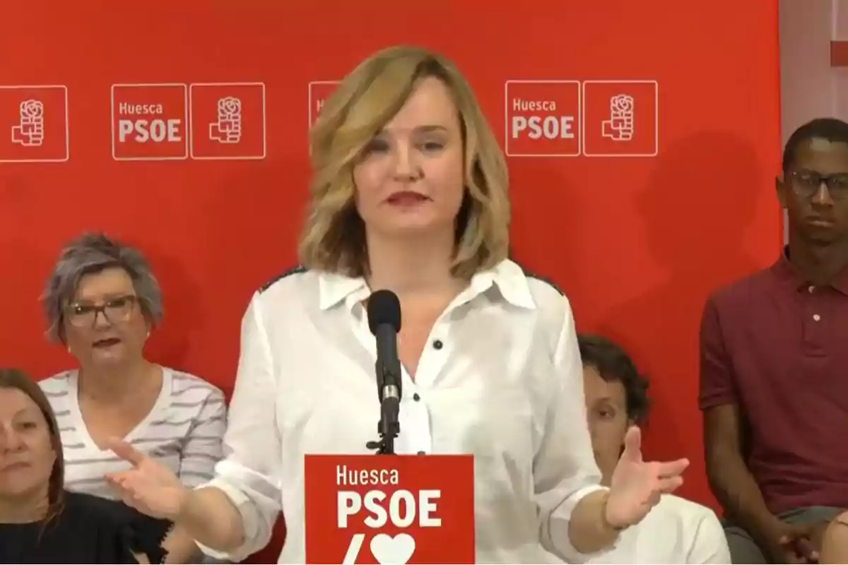 Mujer rubia con camisa blanca hablando en un mitin del PSOE de Huesca con fondo rojo y varias personas sentadas detrás de ella Mujer rubia con camisa blanca hablando en un mitin del PSOE de Huesca con fondo rojo y varias personas sentadas detrás de ella