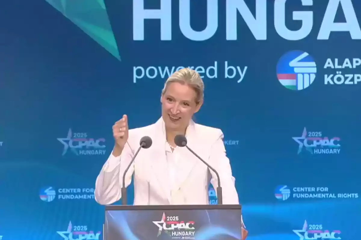 Mujer rubia con saco blanco hablando en un podio durante un evento de CPAC en Hungría con fondo azul y logotipos del evento Mujer rubia con saco blanco hablando en un podio durante un evento de CPAC en Hungría con fondo azul y logotipos del evento