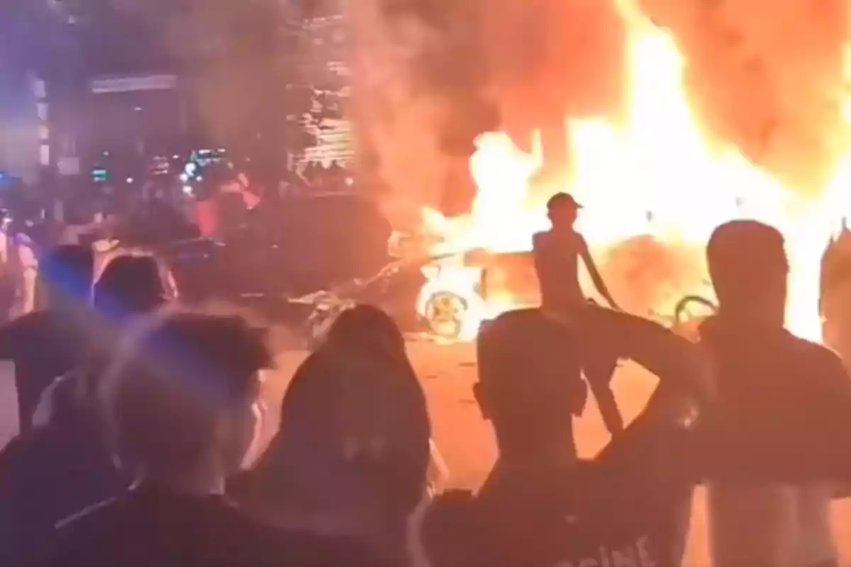 Varias personas observan un automóvil envuelto en llamas durante la noche Varias personas observan un automóvil envuelto en llamas durante la noche