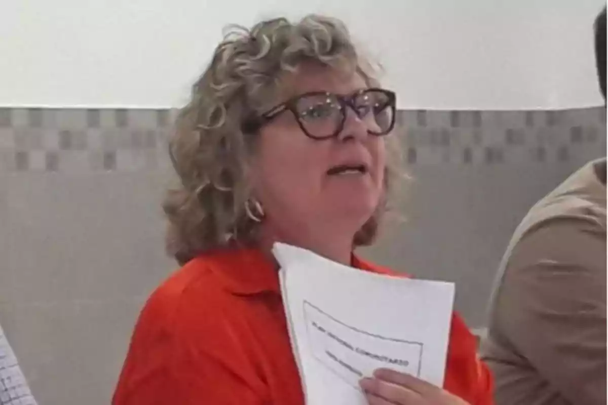 Mujer de cabello rizado y gafas sosteniendo unos papeles mientras habla en un entorno interior Mujer de cabello rizado y gafas sosteniendo unos papeles mientras habla en un entorno interior