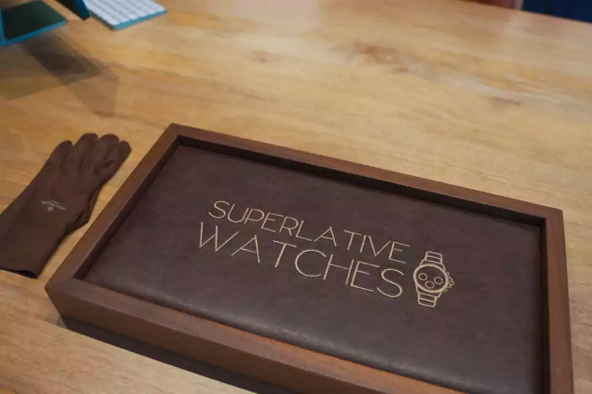 Una caja de madera con una tapa de cuero que tiene grabado "Superlative Watches" junto a un dibujo de un reloj, sobre una mesa de madera clara, con un guante marrón a la izquierda.