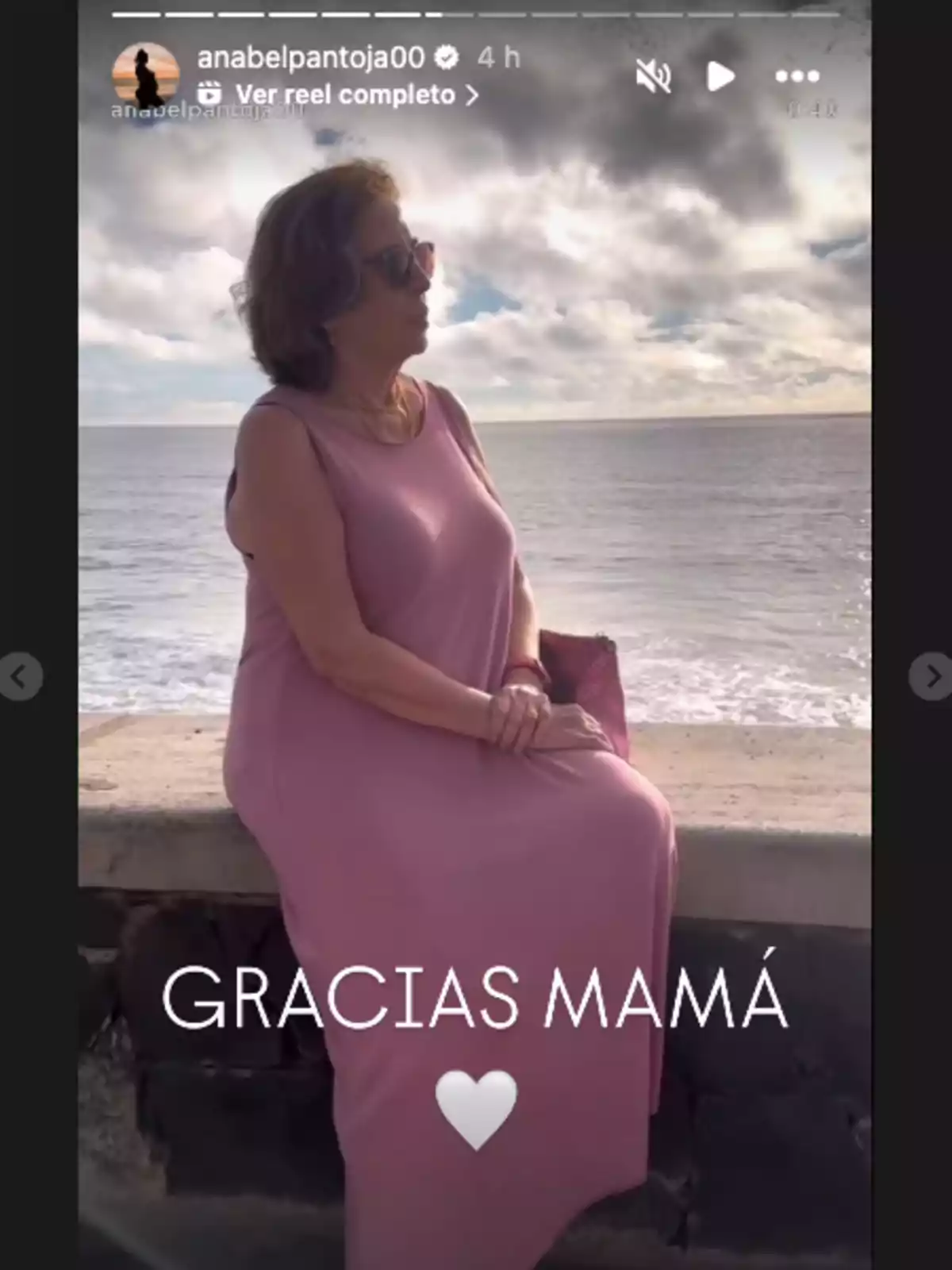 Mercedes Bernal con vestido rosa y gafas de sol está sentada en un muro frente al mar, con un cielo nublado de fondo y el texto Mercedes Bernal con vestido rosa y gafas de sol está sentada en un muro frente al mar, con un cielo nublado de fondo y el texto