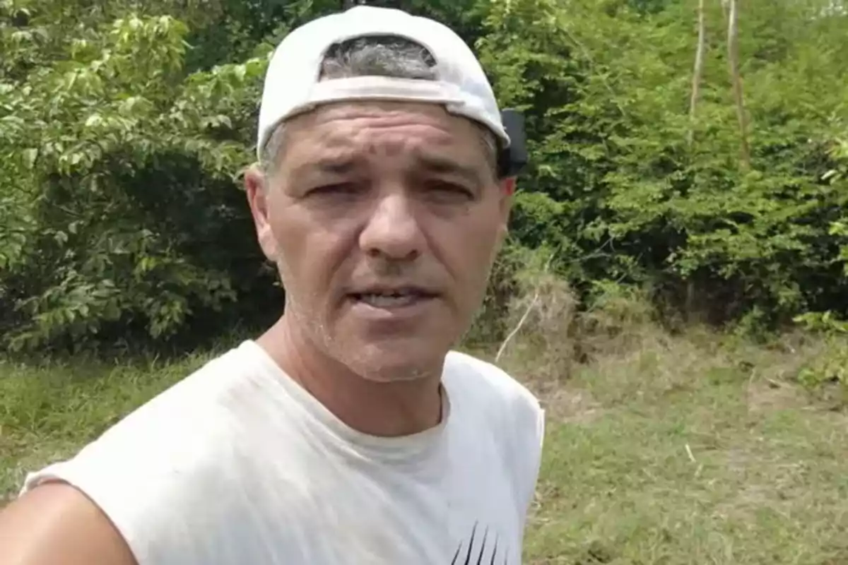 Un hombre con gorra blanca al revés y camiseta blanca está al aire libre rodeado de vegetación. Un hombre con gorra blanca al revés y camiseta blanca está al aire libre rodeado de vegetación.