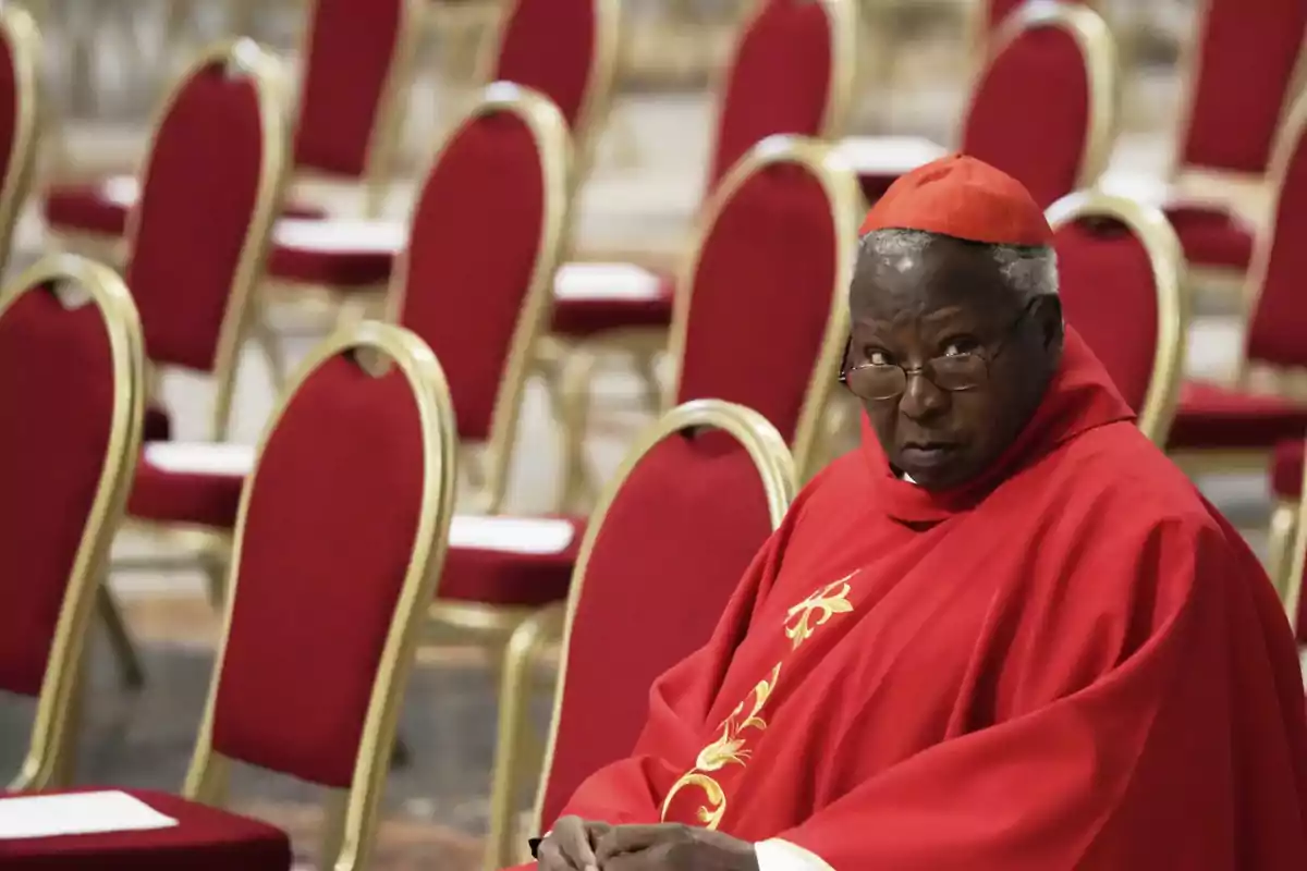 A cardinal in red attire is sitting on a chair in a room with many empty chairs.