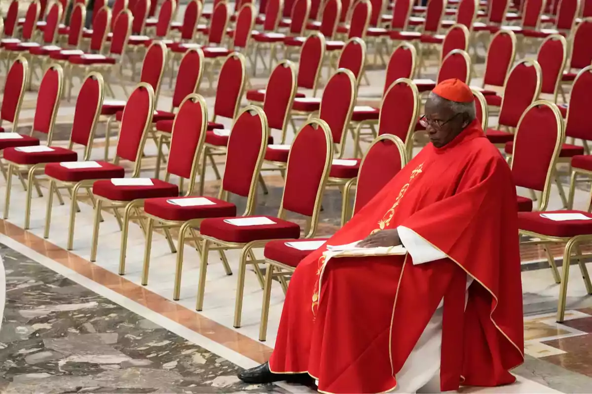 A cardinal dressed in red is sitting in a chair in a room with many empty red chairs. A cardinal dressed in red is sitting in a chair in a room with many empty red chairs.