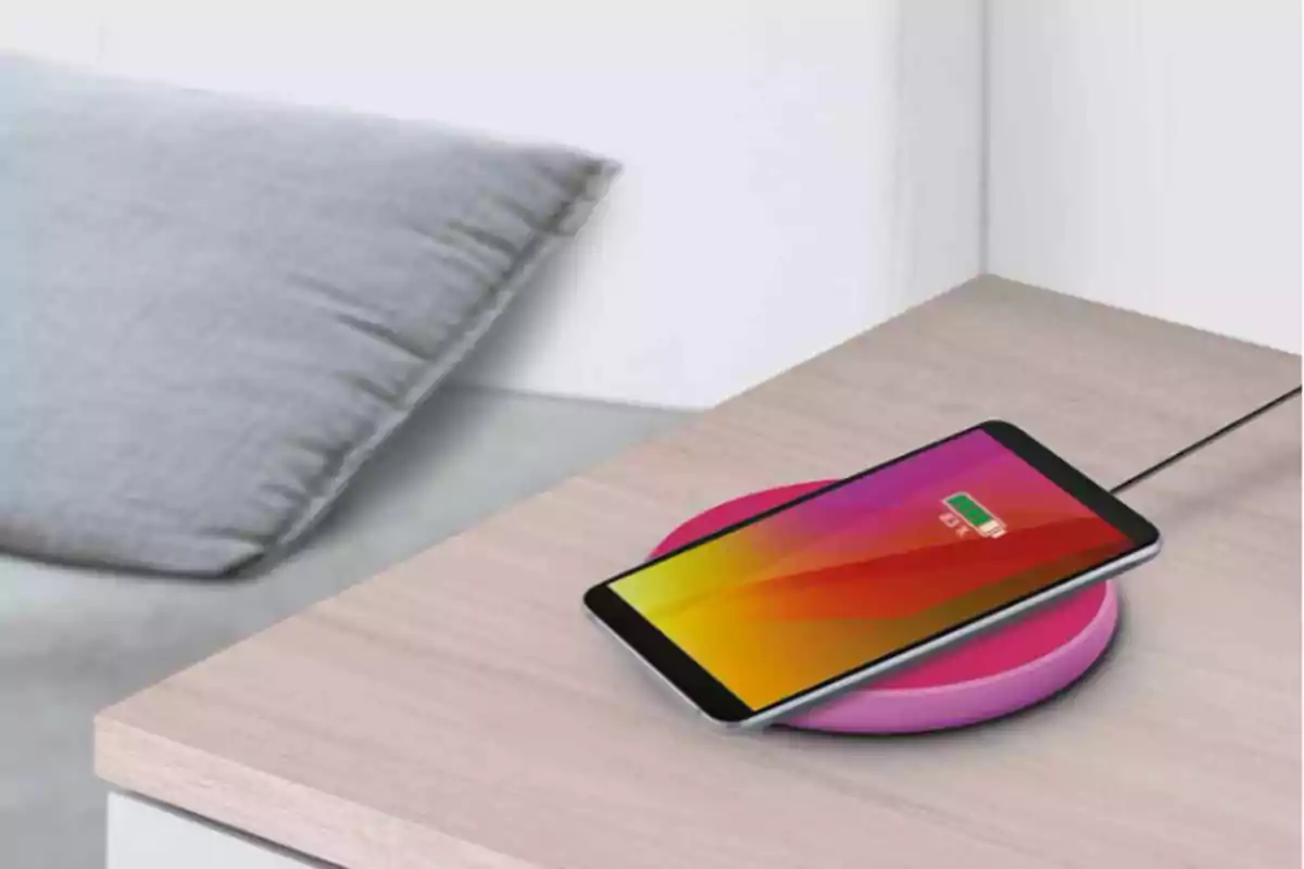 Teléfono inteligente cargando de forma inalámbrica sobre una base rosa en una mesa de madera junto a un cojín gris. Teléfono inteligente cargando de forma inalámbrica sobre una base rosa en una mesa de madera junto a un cojín gris.
