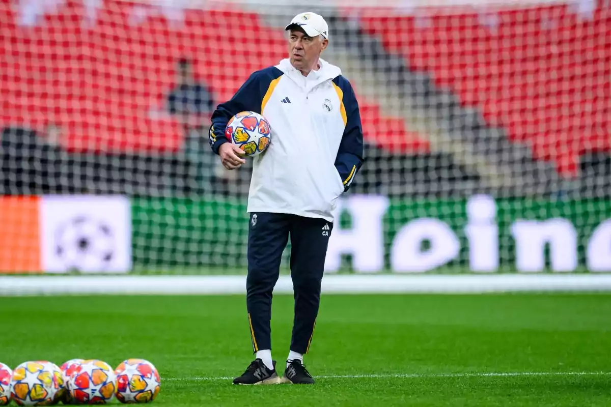 Un entrenador de fútbol con ropa deportiva blanca y azul sostiene un balón en un campo de fútbol con varias pelotas en el suelo y una portería de fondo.