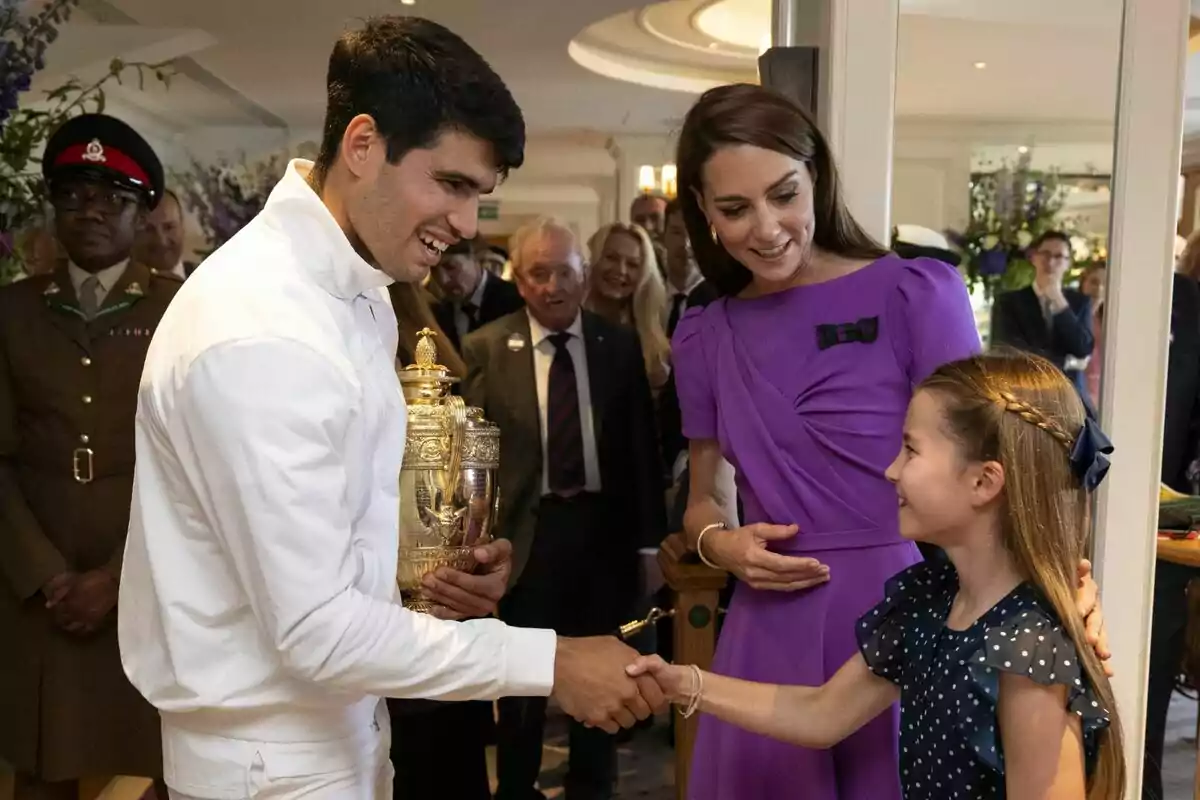 Carlos Alcaraz vestido de blanco sostiene un trofeo mientras estrecha la mano de Charlotte, acompañado por Kate Middleton vestida de morado y varias personas en el fondo. Carlos Alcaraz vestido de blanco sostiene un trofeo mientras estrecha la mano de Charlotte, acompañado por Kate Middleton vestida de morado y varias personas en el fondo.