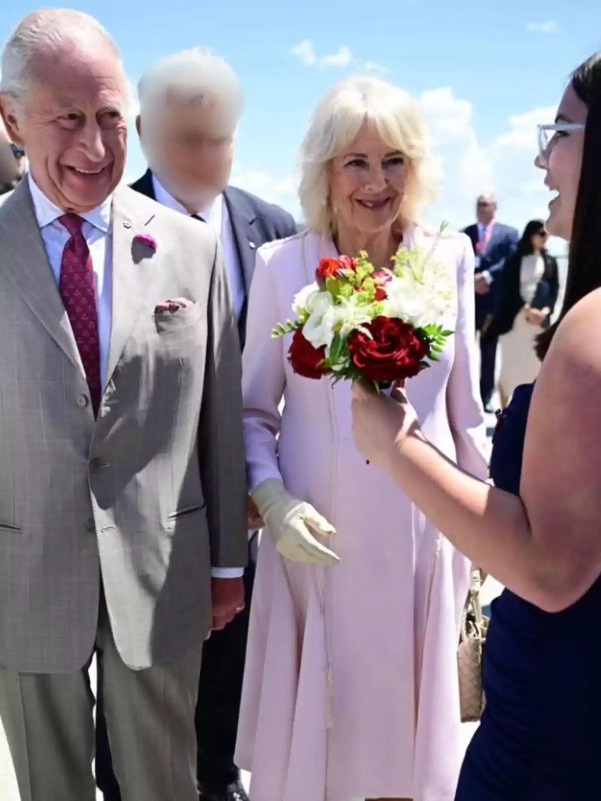 A well-dressed man and woman smile as a young woman hands them a bouquet of red and white flowers at an outdoor event under a blue sky. A well-dressed man and woman smile as a young woman hands them a bouquet of red and white flowers at an outdoor event under a blue sky.