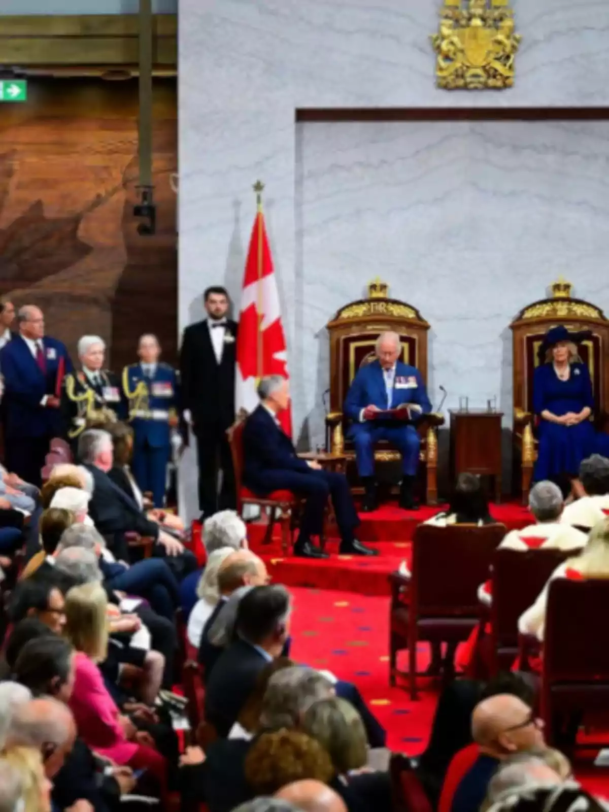 Ceremonia oficial en un salón con alfombra roja, dos personas sentadas en tronos dorados, una bandera de Canadá y público observando Ceremonia oficial en un salón con alfombra roja, dos personas sentadas en tronos dorados, una bandera de Canadá y público observando