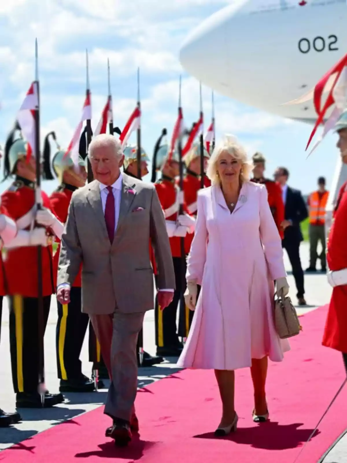 An elegantly dressed couple walks down a red carpet while soldiers in dress uniforms and carrying spears form a guard of honor next to a white airplane. An elegantly dressed couple walks down a red carpet while soldiers in dress uniforms and carrying spears form a guard of honor next to a white airplane.