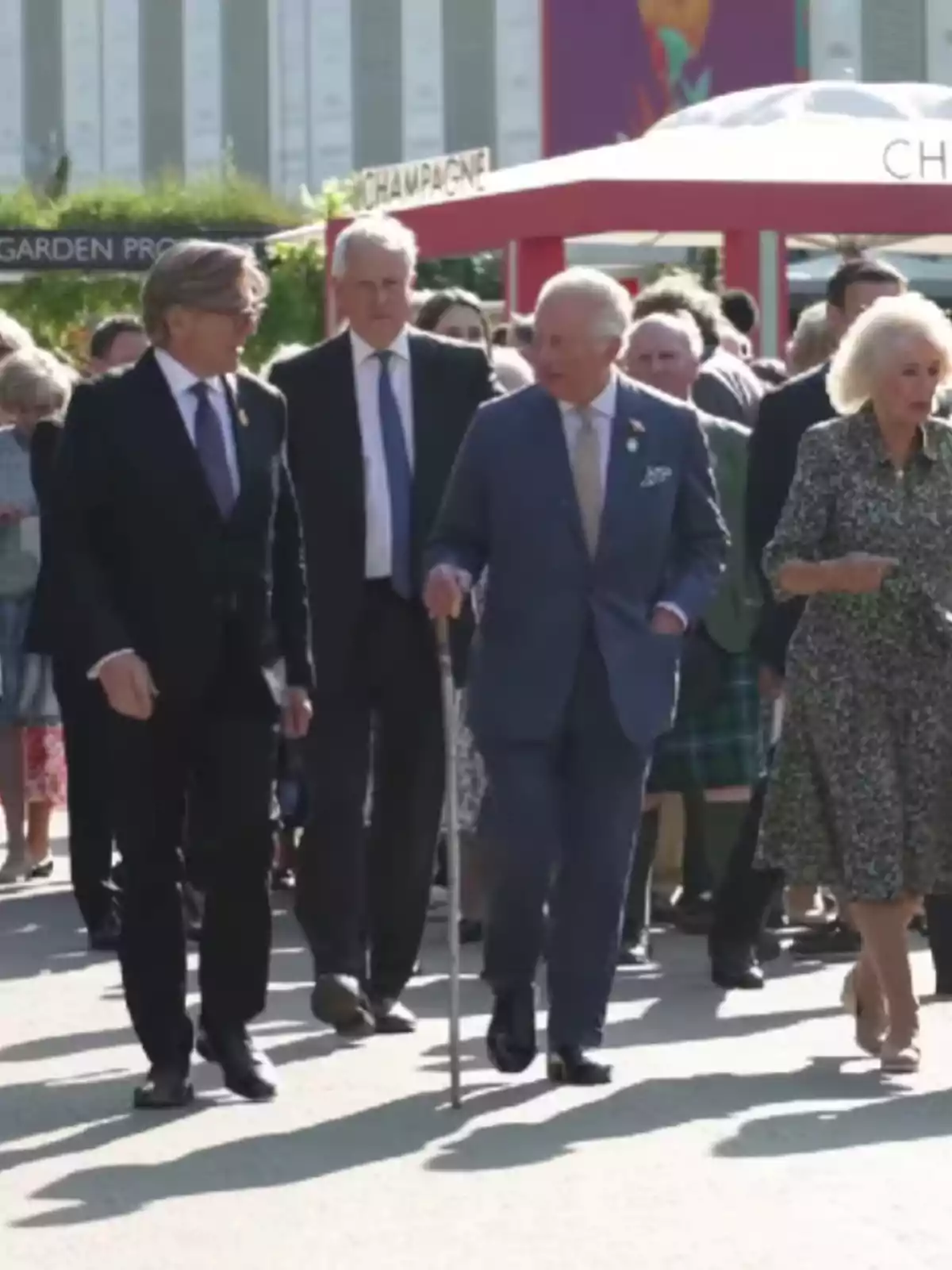 Un grupo de personas caminando al aire libre en un evento, con un hombre mayor usando un bastón y una mujer con un vestido estampado. Un grupo de personas caminando al aire libre en un evento, con un hombre mayor usando un bastón y una mujer con un vestido estampado.