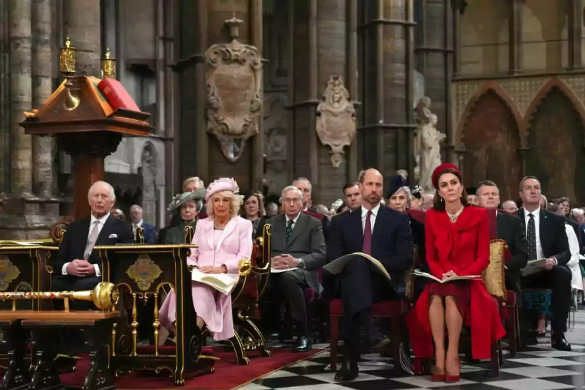 A group of elegantly dressed people is seated at a formal event inside a historic building with detailed architecture.